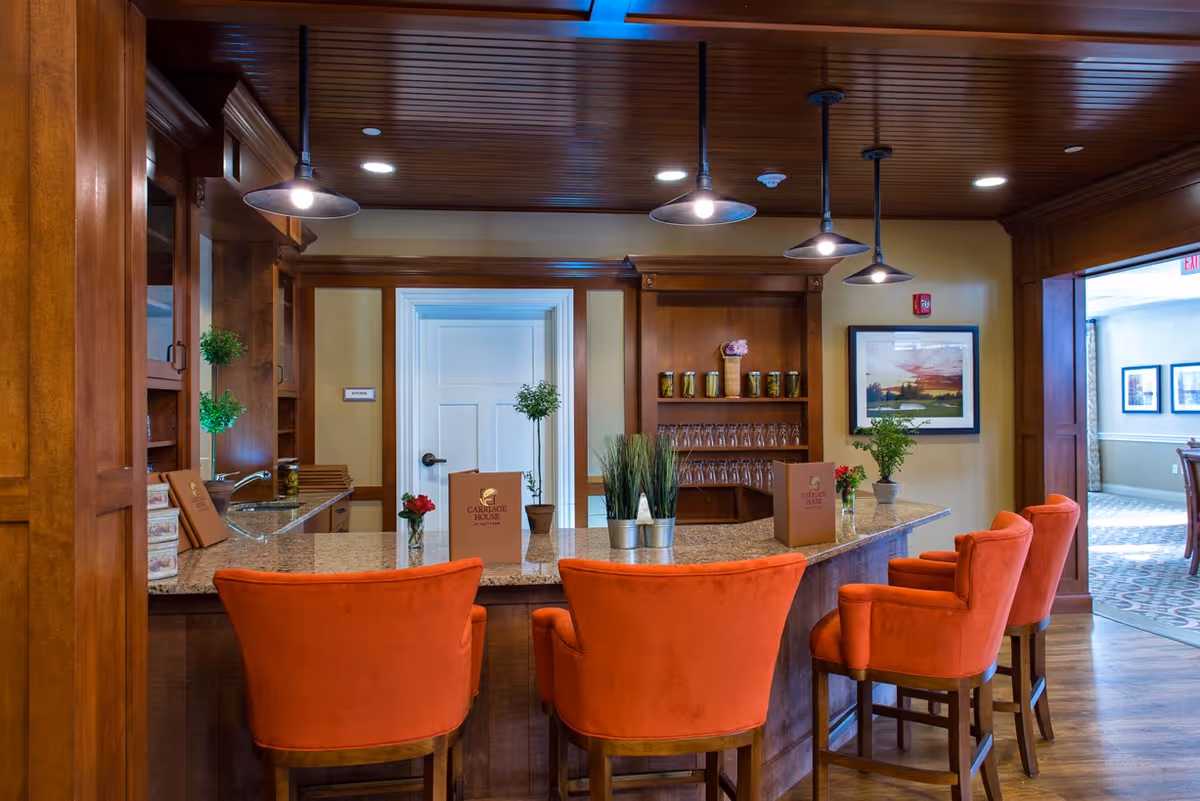Interior view of a senior living facility's bar or lounge area with a granite countertop, four orange upholstered bar stools, wooden cabinetry, pendant lighting, and decorative plants. There are menus labeled 'Carriage House' on the counter and framed artwork on the wall.