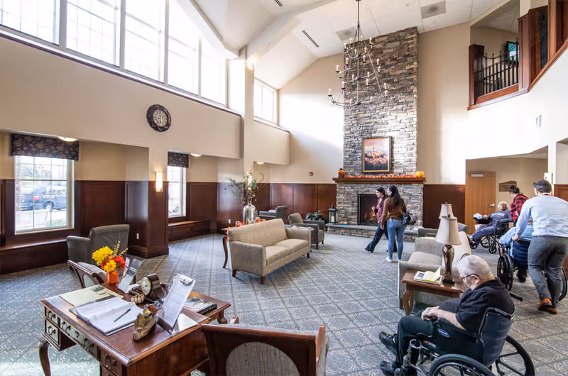 Spacious two-story common lounge with a large stone fireplace, sofas and chairs, a chandelier, and residents in wheelchairs.