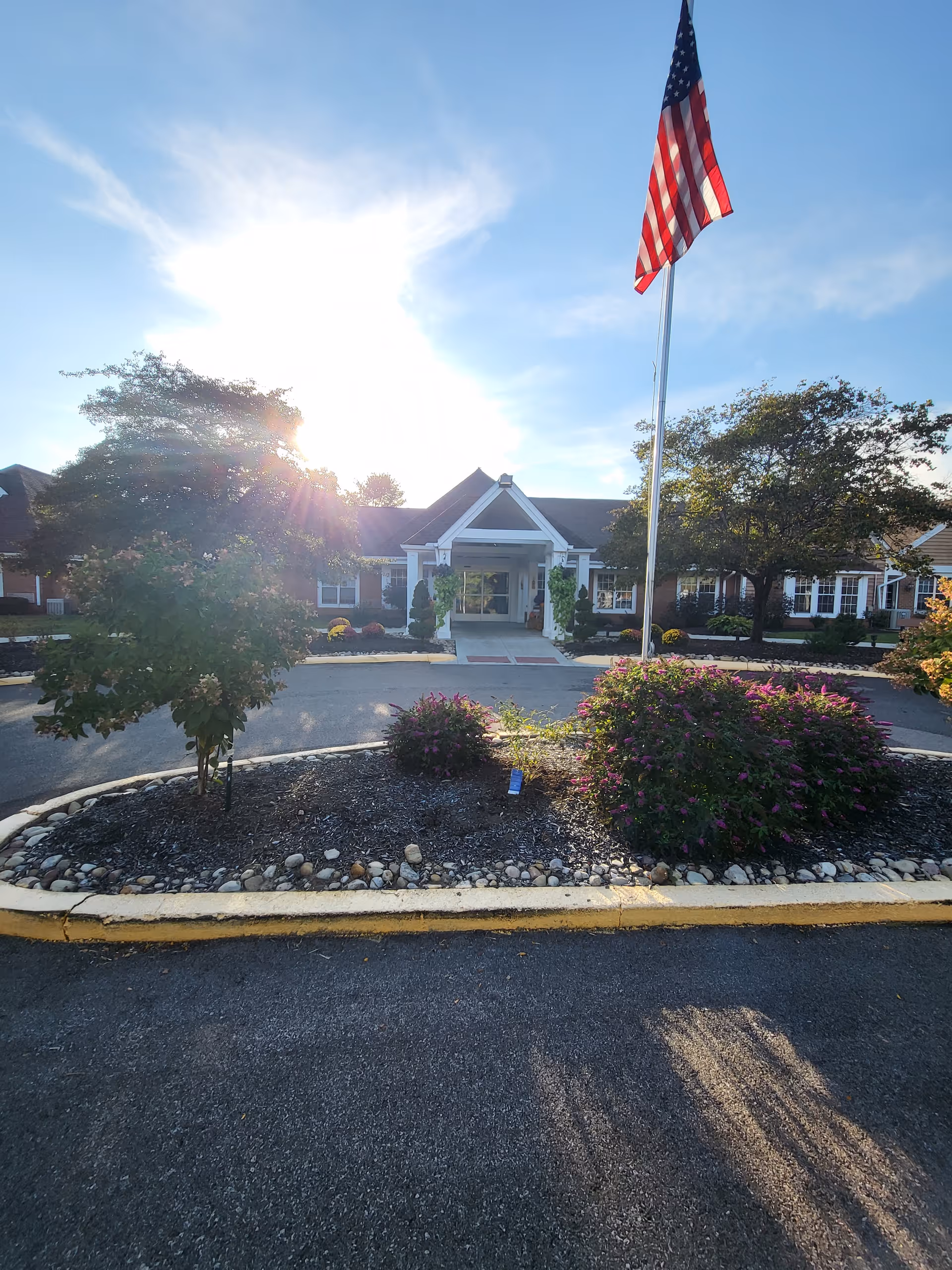 Front exterior view of Phoenix of Fairlawn (formerly known as Windsong) senior living facility with a circular driveway, landscaped garden bed with bushes and flowers, an American flag on a tall flagpole, and trees on either side under a bright sky with the sun shining.