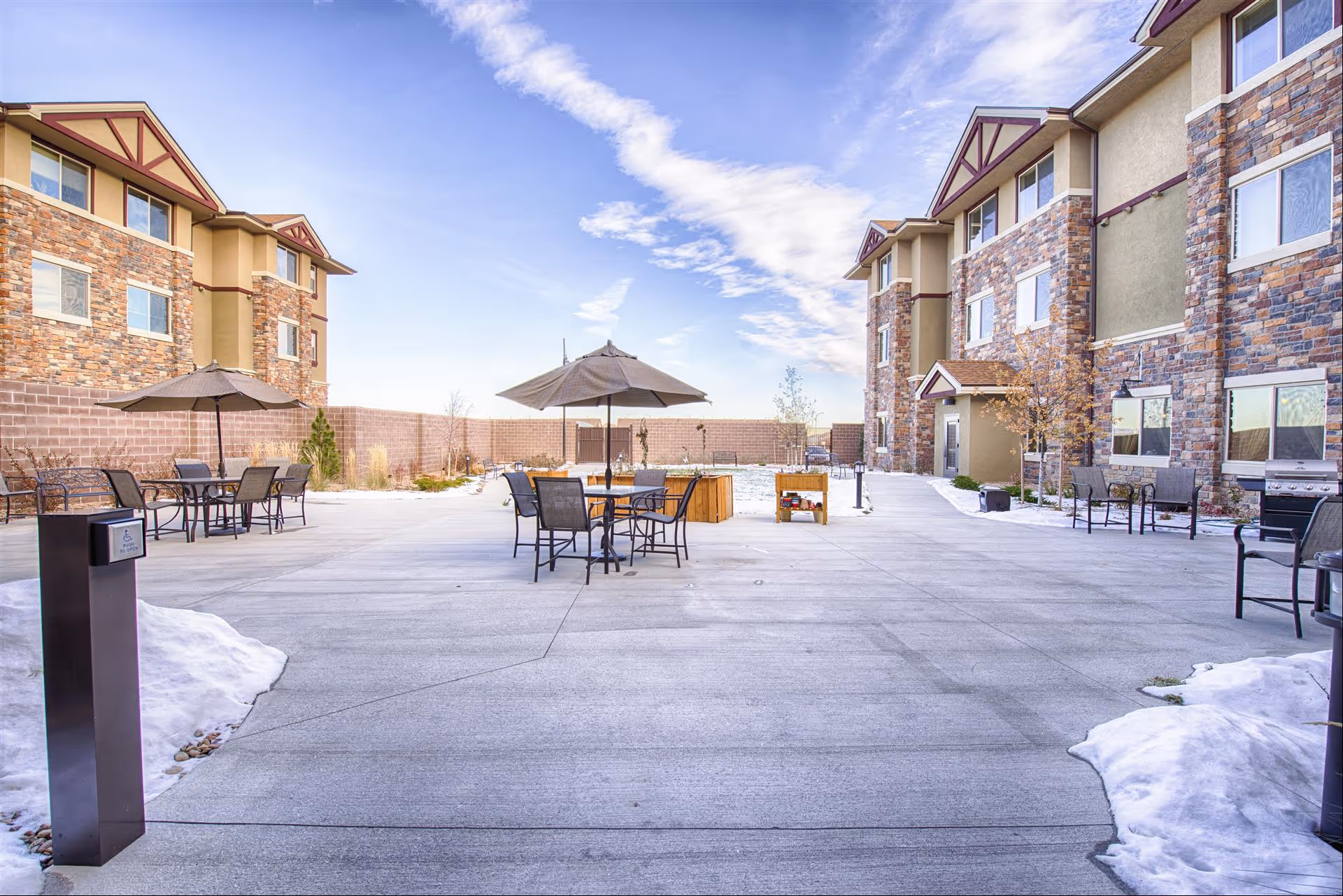 Outdoor courtyard area of a senior living facility with concrete pavement, several tables with chairs and umbrellas, surrounded by multi-story buildings with stone and beige exterior walls. There is some snow on the ground and a clear blue sky with clouds above.