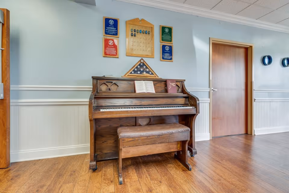 An upright wooden piano with a matching bench in front of a light blue wall. On the piano are two open books, and above it is a folded American flag in a triangular display case surrounded by plaques and awards. To the right is a closed wooden door and two decorative wall-mounted plates. The floor is wooden.