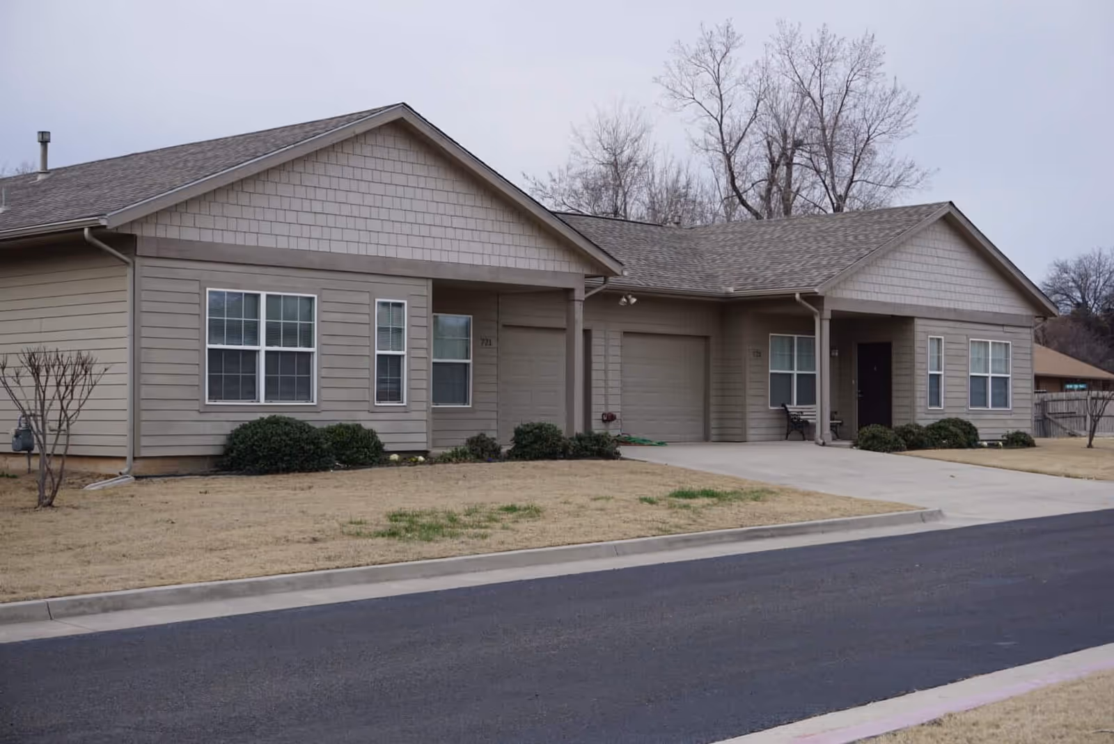 Exterior view of a single-story residential building with beige siding and two attached garages. The building has multiple windows and small bushes in front, with a paved driveway and road in the foreground. Leafless trees are visible in the background under an overcast sky.