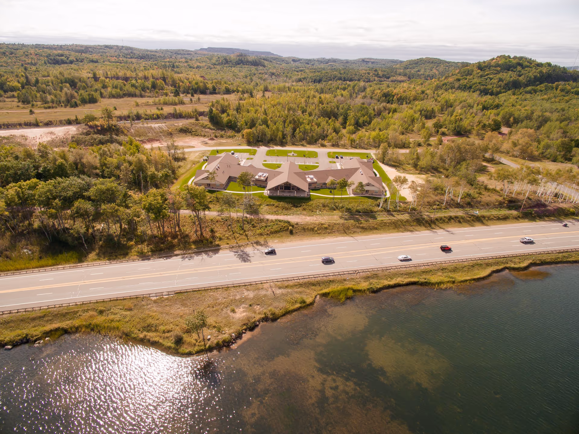 Aerial view of a single-story lakeside senior living building set behind a highway, surrounded by trees and rolling hills.