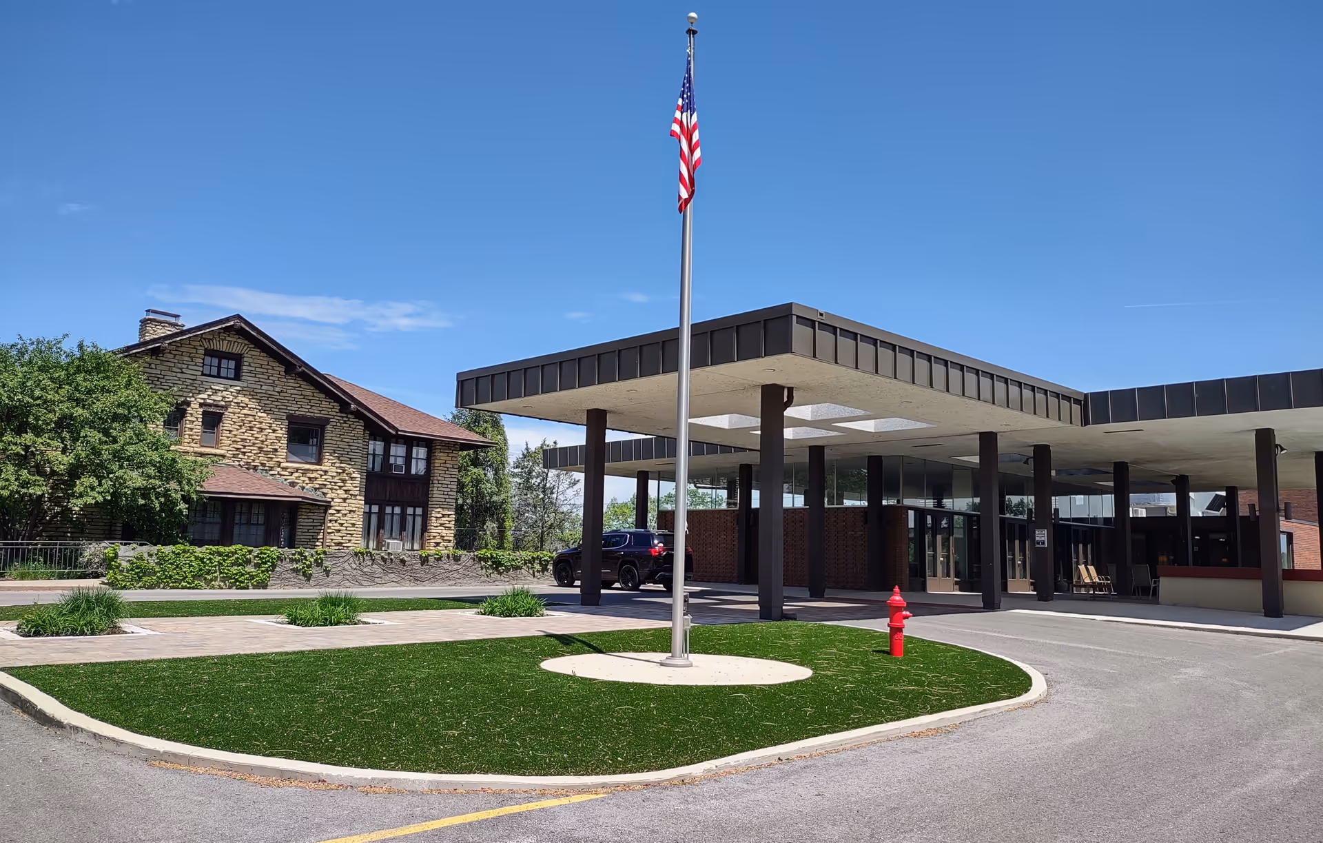 Front porte-cochère entrance of a retirement community with an American flag on a central pole, a stone building to the left, a parked car, and a grassy island with a fire hydrant.