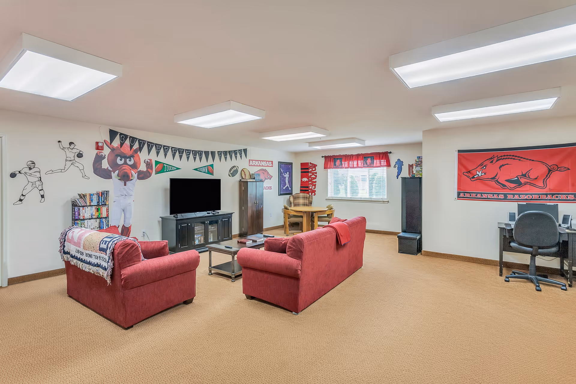A spacious lounge with red sofas facing a TV, a game table, desk, and Arkansas Razorbacks decorations on the walls.