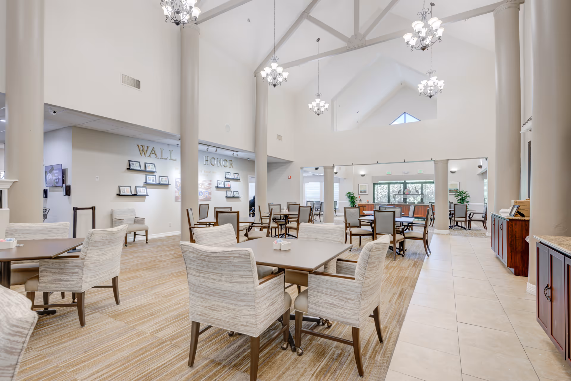Spacious dining area in The Continental Retirement Community with multiple tables and cushioned chairs arranged neatly. The room features high vaulted ceilings with chandeliers, large columns, and a wall labeled 'Wall of Honor' displaying framed photos. The floor is a combination of carpet and tile, and there are large windows letting in natural light.