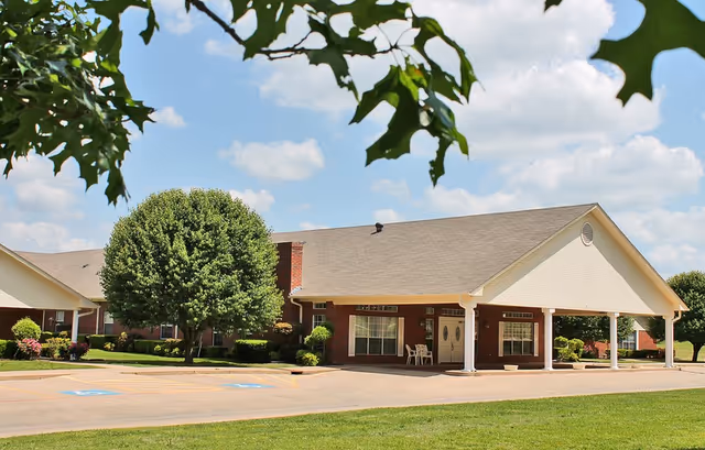 Exterior view of a single-story brick building with a covered entrance supported by white columns. The building is surrounded by green grass, trees, and shrubs under a partly cloudy blue sky.