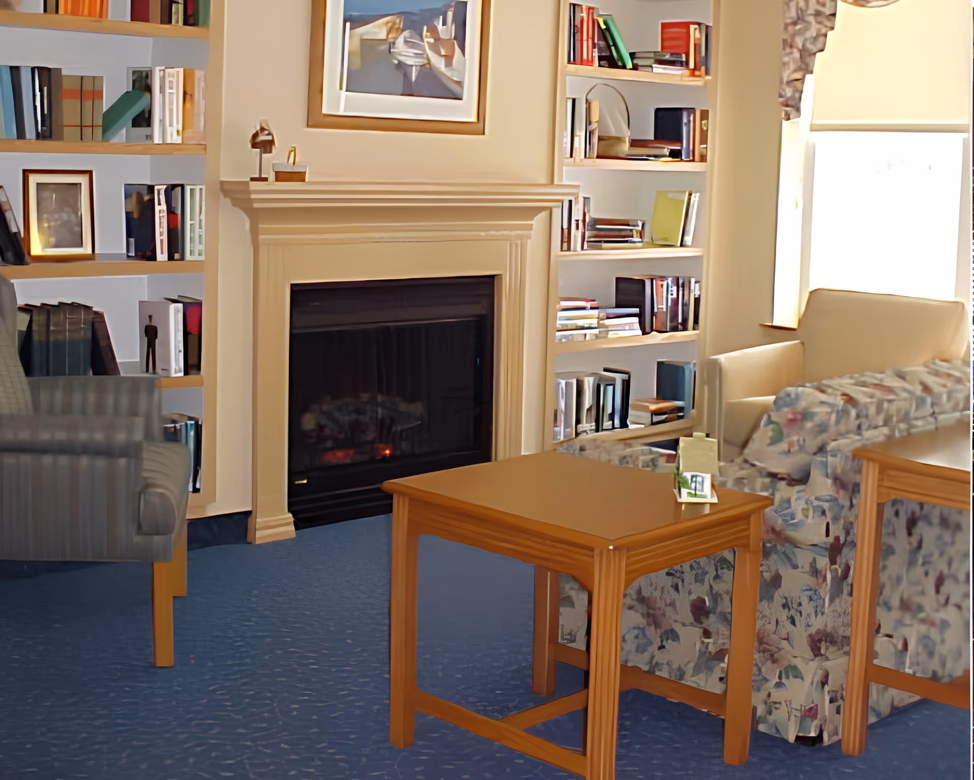 A cozy living room area with a fireplace in the center, flanked by built-in bookshelves filled with books and decorative items. There are upholstered chairs and a floral-patterned sofa around a wooden table. A window with a light-colored blind allows natural light into the room.