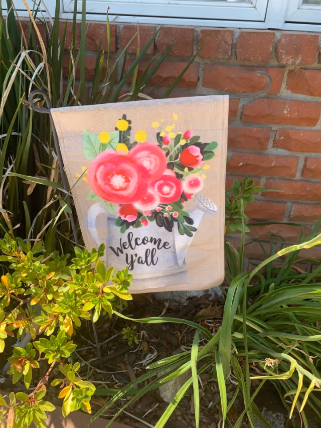 A decorative garden flag with a painted watering can holding bright pink and red flowers and green leaves. The flag has the text 'Welcome y'all' and is placed in a garden bed with green plants and a brick wall in the background.