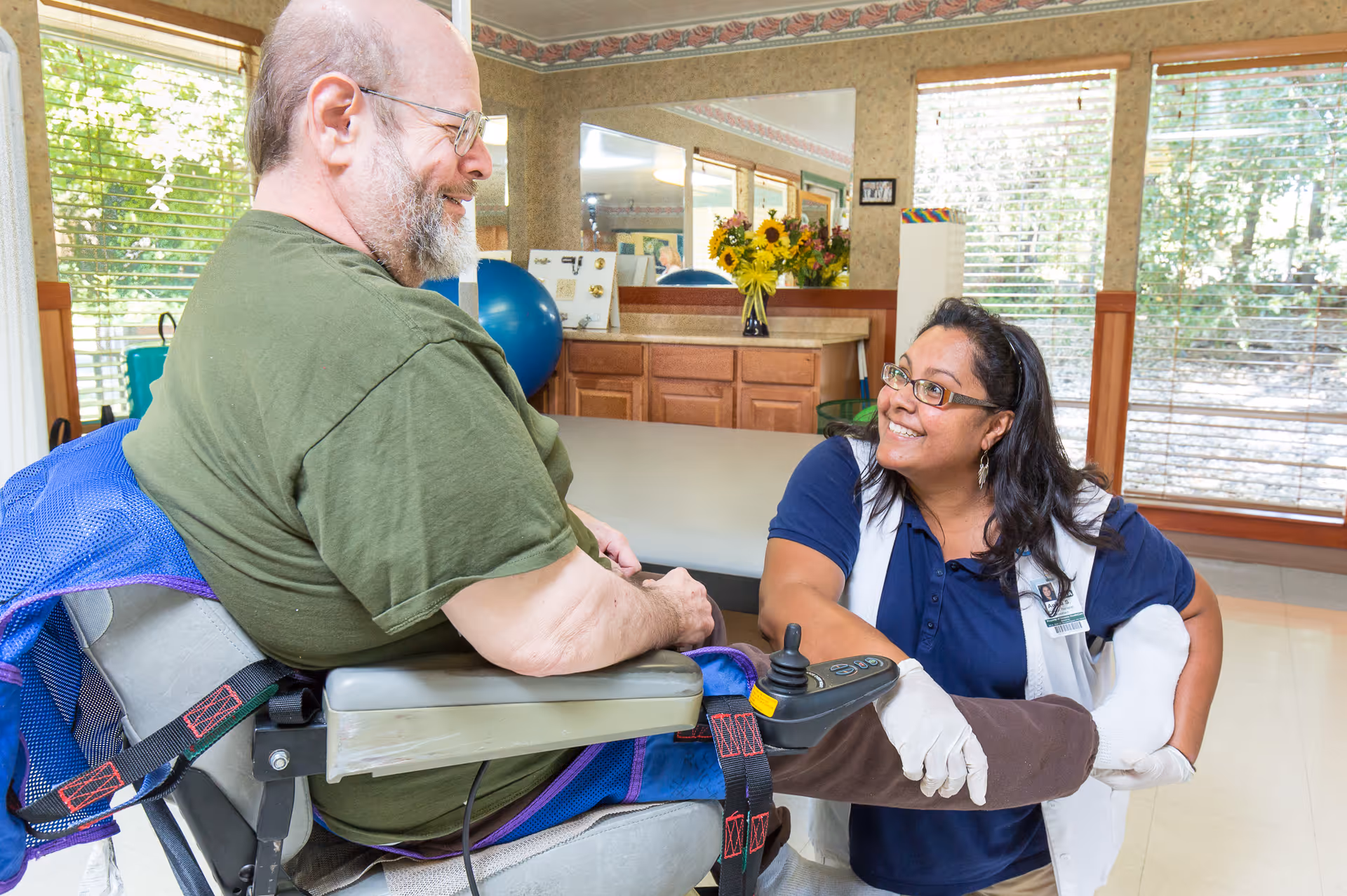A smiling female caregiver wearing gloves is assisting a male resident in a wheelchair by holding and stretching his leg in a bright room with large windows and wooden blinds.