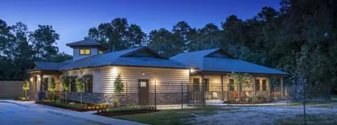 Dusk view of a single-story senior living facility with lit exterior and fenced front.