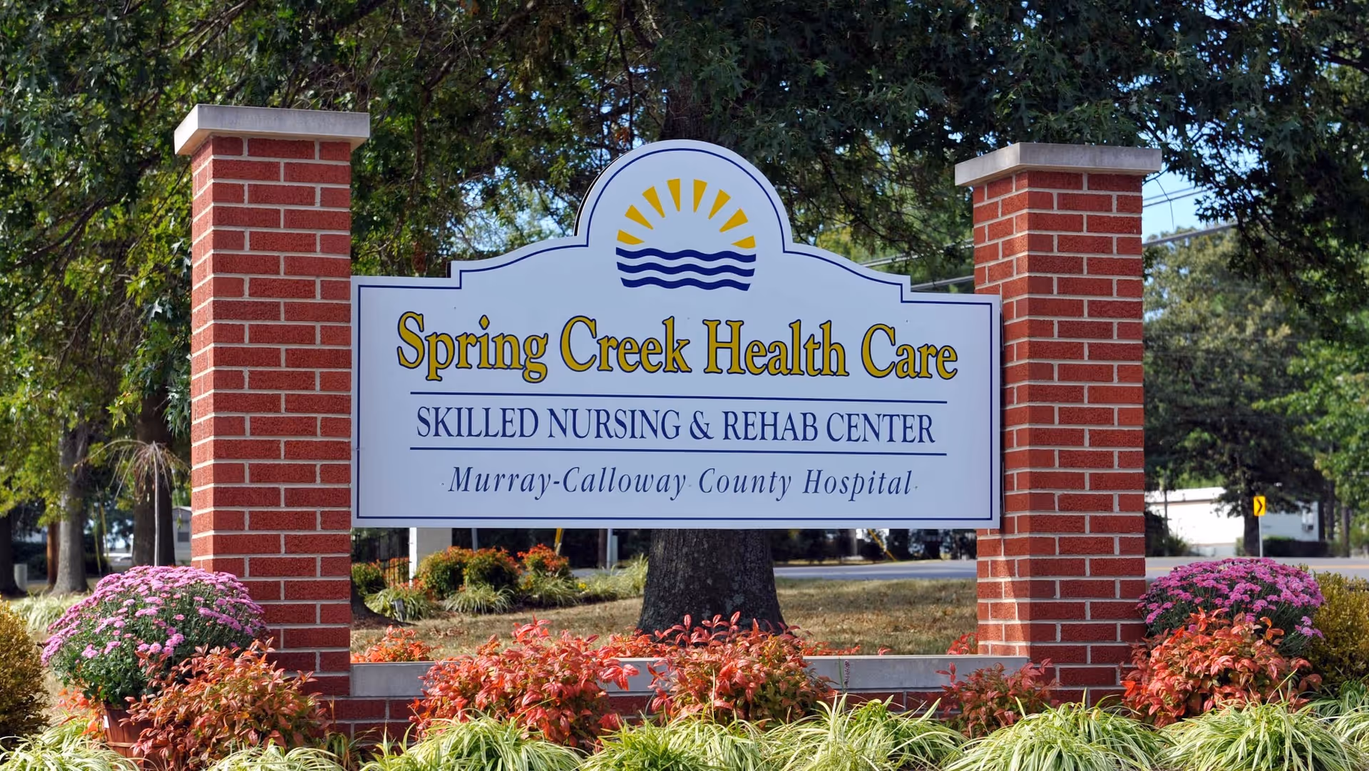 Outdoor sign for Spring Creek Health Care Skilled Nursing & Rehab Center, part of Murray-Calloway County Hospital, mounted between two red brick pillars with landscaping including flowers and bushes around the base.