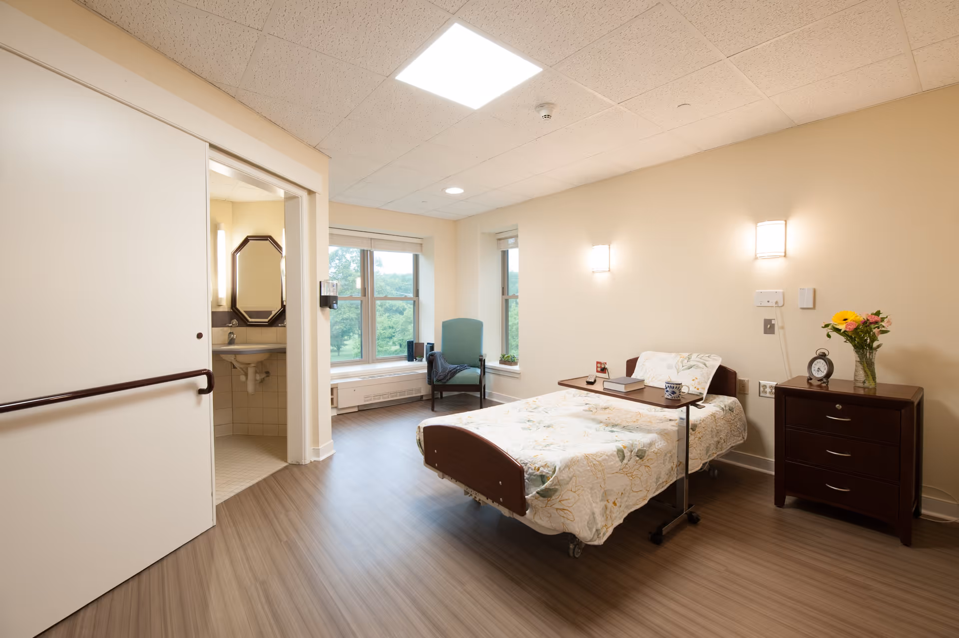A bright and clean senior living bedroom with a single bed covered in a floral quilt, a bedside table with a vase of flowers and an alarm clock, a small overbed table with a book and a mug, a green armchair near two large windows, and an open door leading to a bathroom with a sink and mirror.
