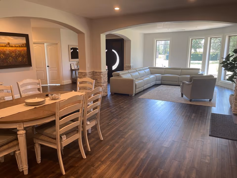Open-plan living area with a dining table on the left and a beige sectional sofa and armchair by large windows.