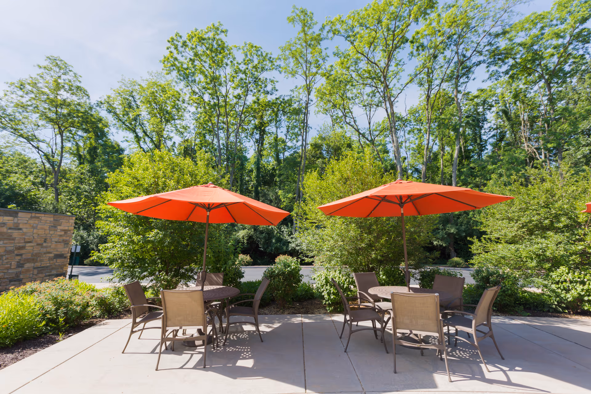 Outdoor patio area with two round tables, each shaded by a large orange umbrella, surrounded by six brown chairs. The patio is bordered by green bushes and tall trees under a clear blue sky.