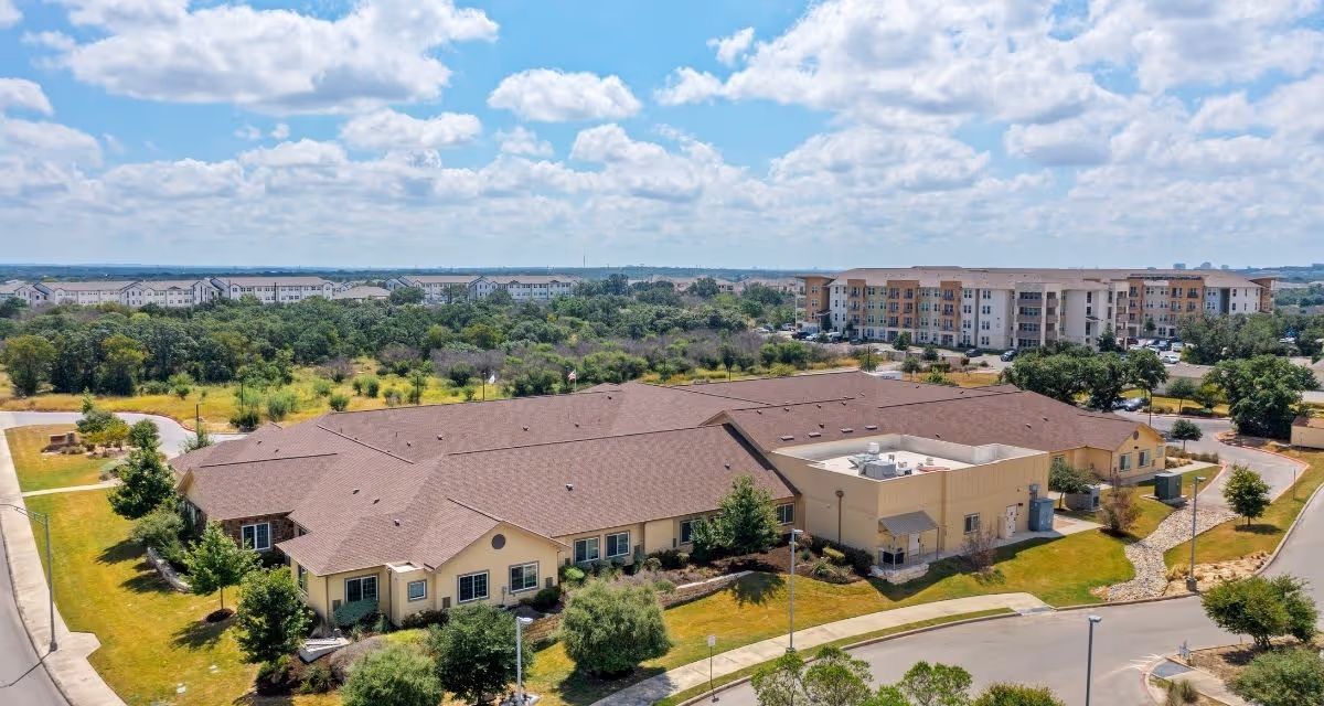 Aerial view of a single-story memory care building surrounded by lawns and trees with an apartment complex and cloudy sky in the background.