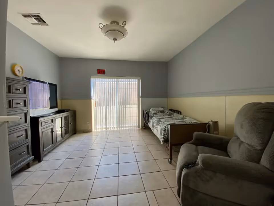 A simple room with tiled floor, a single bed with a patterned blanket, a large cushioned recliner chair, and a wooden entertainment center with a flat-screen TV. There is a sliding glass door with vertical blinds letting in natural light, and an exit sign above the door. The walls are painted gray on the upper half and beige on the lower half.