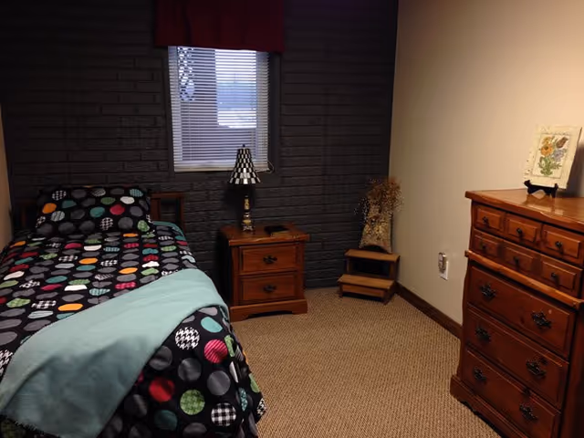 A small bedroom with a single bed covered in a black comforter with colorful circular patterns and a light blue blanket at the foot. Next to the bed is a wooden nightstand with a lamp featuring a black and white checkered shade. The wall behind the bed is dark gray brick with a small window covered by blinds and a red valance. On the right side of the room is a wooden dresser with multiple drawers and a framed floral picture on top. The floor is carpeted in a light brown color.