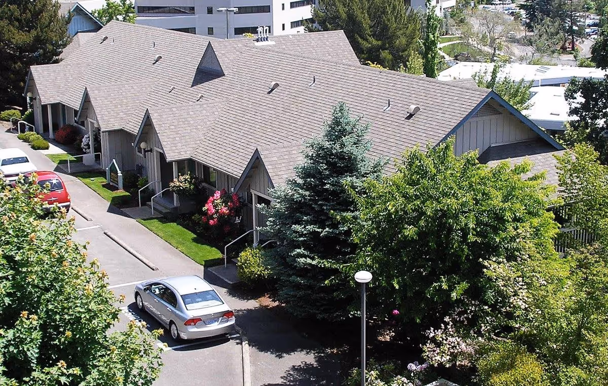 Aerial view of a single-story housing complex with pitched roofs, landscaped trees and shrubs, and cars parked along a driveway.