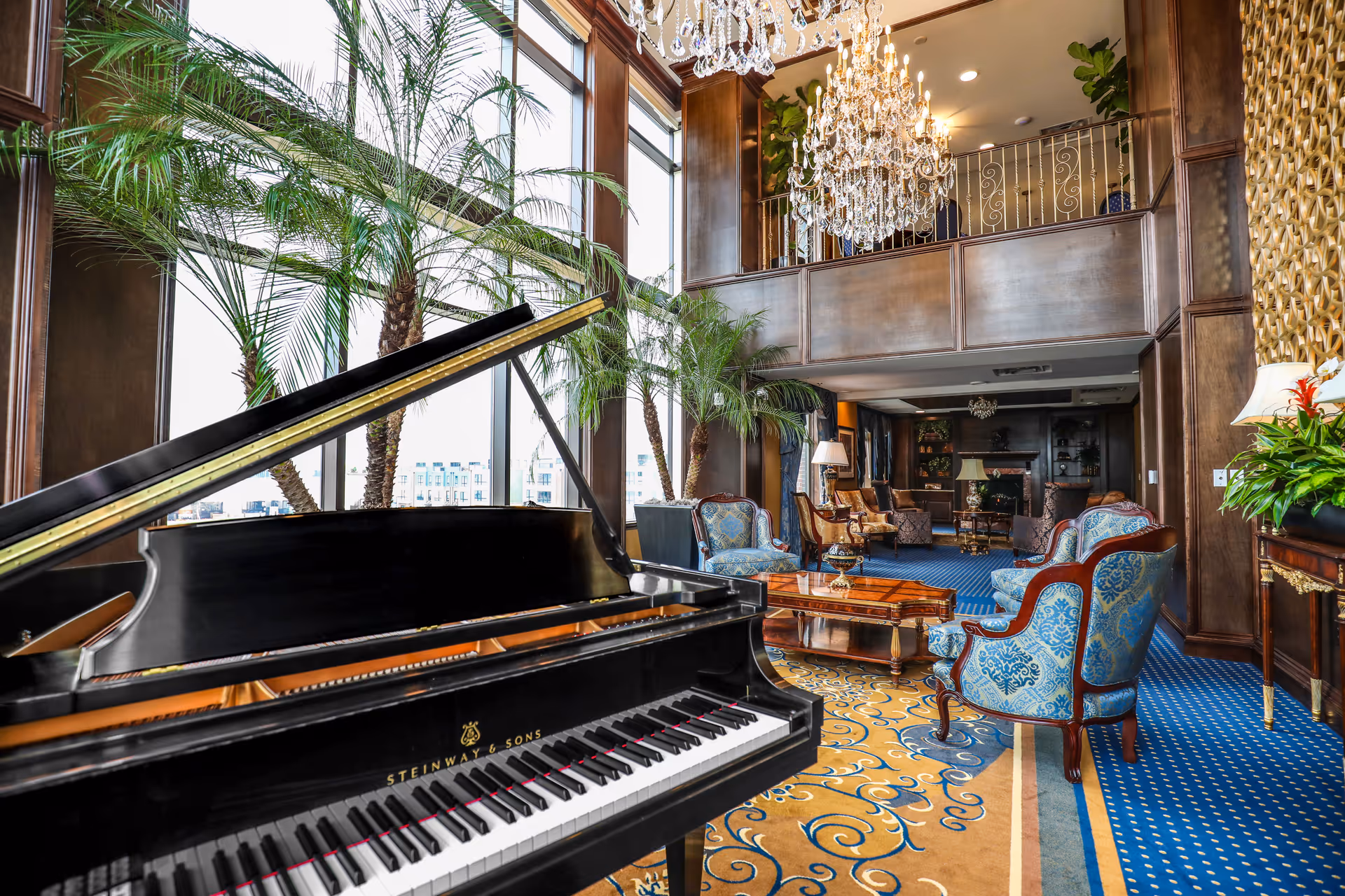 Elegant senior living facility common area with a black Steinway & Sons grand piano in the foreground, large windows with tall palm plants, blue and gold patterned carpet, ornate wooden furniture with blue upholstery, a crystal chandelier hanging from the ceiling, and a cozy seating area with lamps and a fireplace in the background.