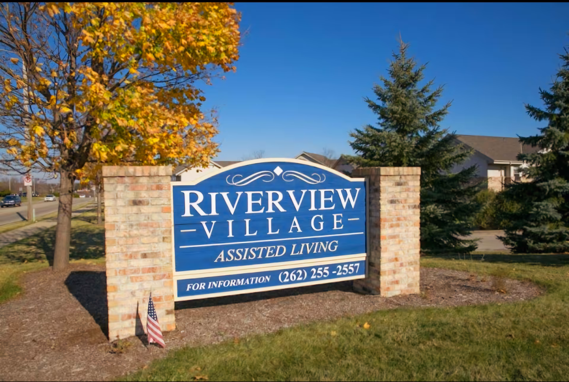 Outdoor view of a large blue and white sign for Riverview Village Assisted Living, mounted between two brick pillars, with trees and a clear blue sky in the background.