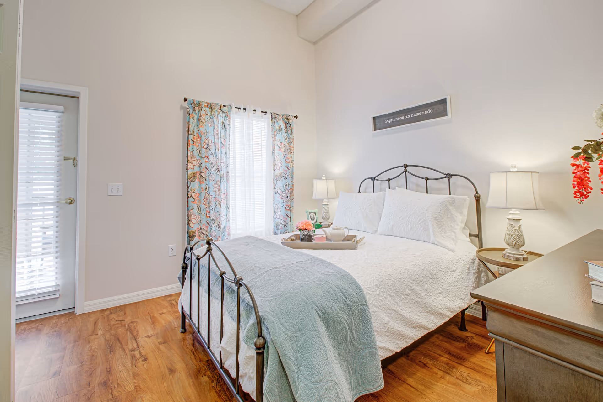 A bright bedroom with a metal bed frame, white bedding, and a light blue quilt at the foot of the bed. There are two bedside tables with lamps, a window with floral curtains, and a door with blinds. A framed sign above the bed reads 'happiness is homemade'.