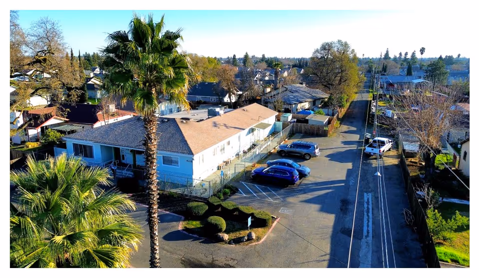 Aerial view of a single-story Lakewood Villa building with palm trees, a parking lot with cars, and surrounding neighborhood homes.