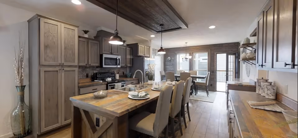 A spacious kitchen and dining area featuring wooden cabinetry and countertops, a kitchen island with seating for four, stainless steel appliances including a microwave and stove, pendant lighting above the island, and a dining table with chairs near large windows and a glass door leading outside.
