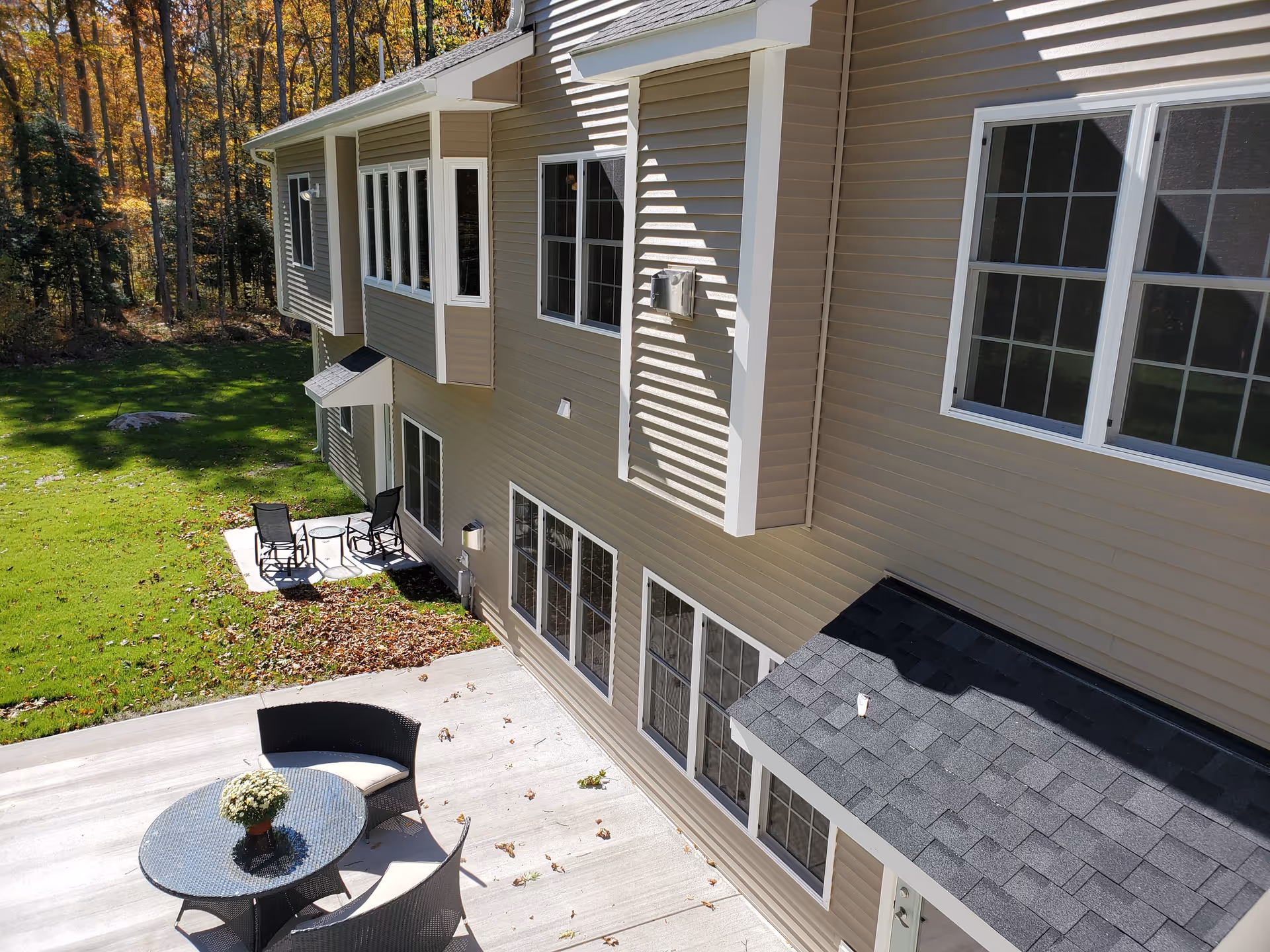 Exterior view of a beige residential building with multiple windows, a patio area with a round glass table and chairs, and a small seating area with two chairs and a table on a grassy lawn surrounded by trees with autumn foliage.