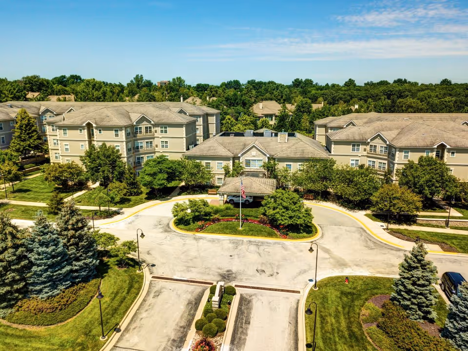 Aerial view of Town Village of Leawood senior living facility showing multiple beige residential buildings surrounded by green trees and landscaped lawns. A circular driveway with an American flag in the center is visible in front of the main building under a clear blue sky.