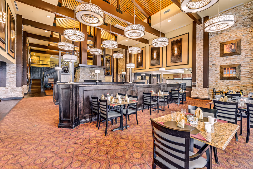 Spacious dining room with multiple tables set with napkins, cups, and glasses. The room features high ceilings with modern circular pendant lights, stone accent walls, and warm earth-tone decor. There are dark wooden partitions separating seating areas and framed artwork on the walls.