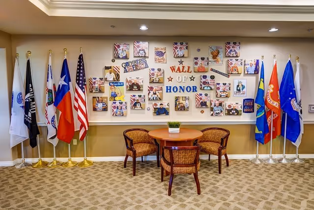 A seating area with a round wooden table and three upholstered chairs in front of a wall decorated with a 'Wall of Honor' display featuring patriotic images and messages. The wall is flanked by multiple flags representing various organizations and states.
