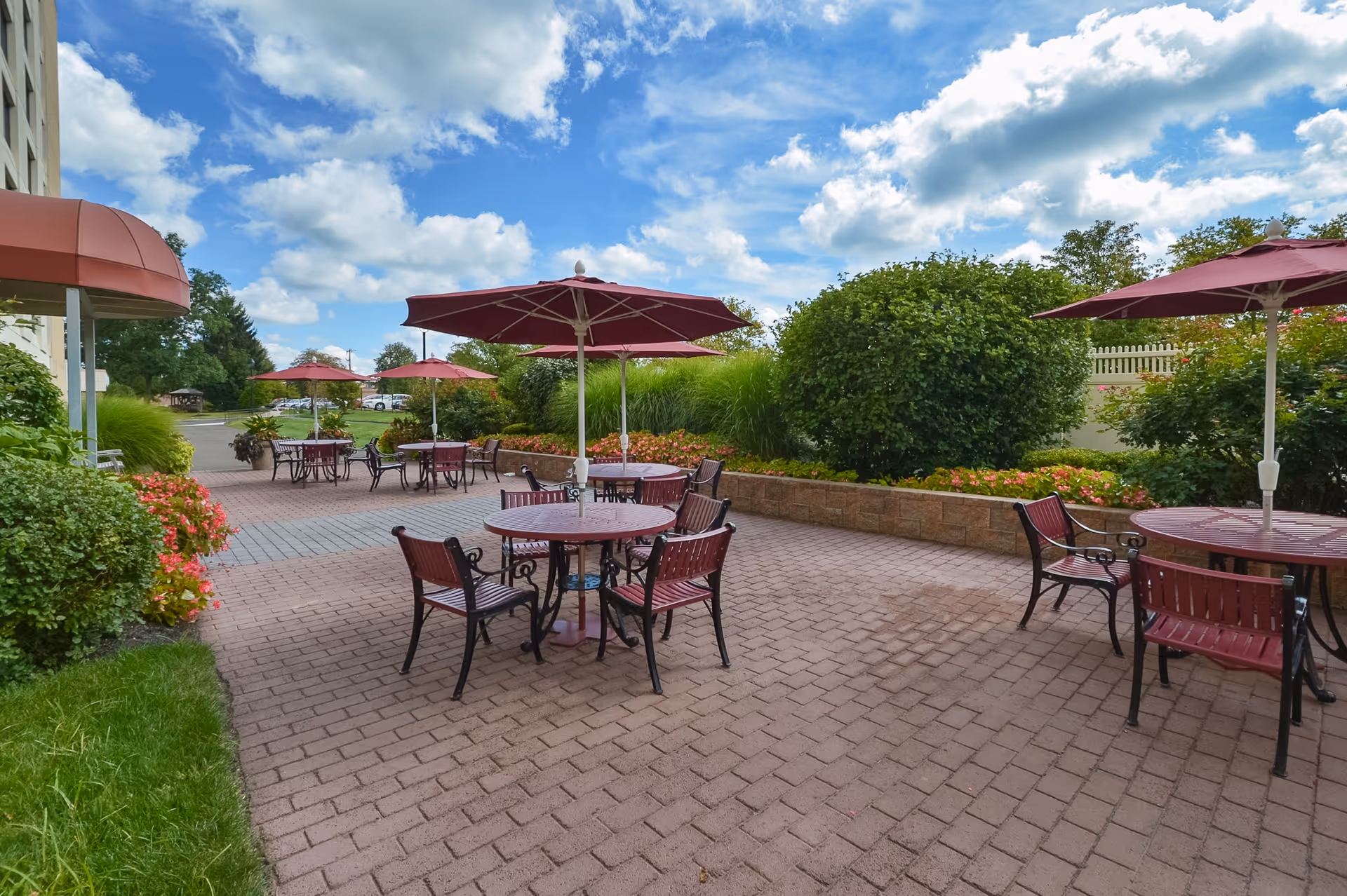 Outdoor patio area with several round tables and chairs, each table shaded by a red umbrella. The patio is paved with bricks and surrounded by green bushes and plants under a partly cloudy blue sky.