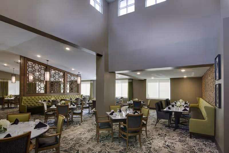 A spacious and well-lit dining room with multiple tables set for meals. The room features green cushioned bench seating along the walls, wooden chairs with dark cushions, and tables adorned with white flowers and neatly folded black napkins. Large windows with green curtains allow natural light to fill the space, and decorative wooden panels add an elegant touch to the room.