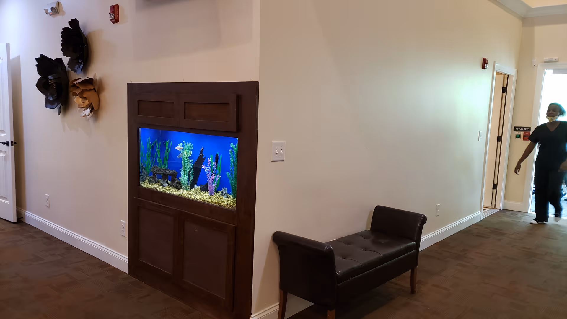 Interior hallway of a senior care home with a built-in fish tank embedded in a wooden panel on the wall. There is a dark brown cushioned bench against the wall and decorative wall art featuring large flower shapes. A person wearing dark clothing and a face mask is walking in the hallway near an exit door.