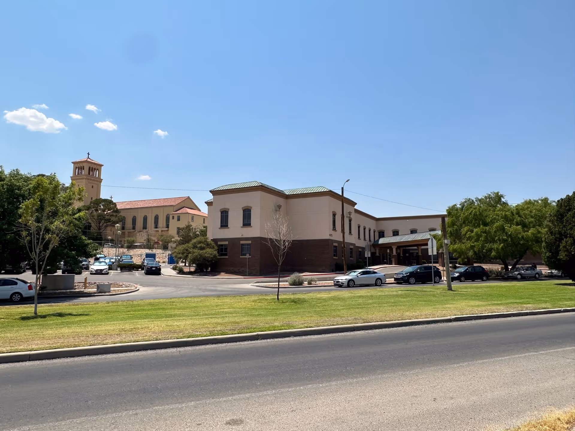 Two-story care facility building with parked cars, a grassy lawn and trees under a clear blue sky.