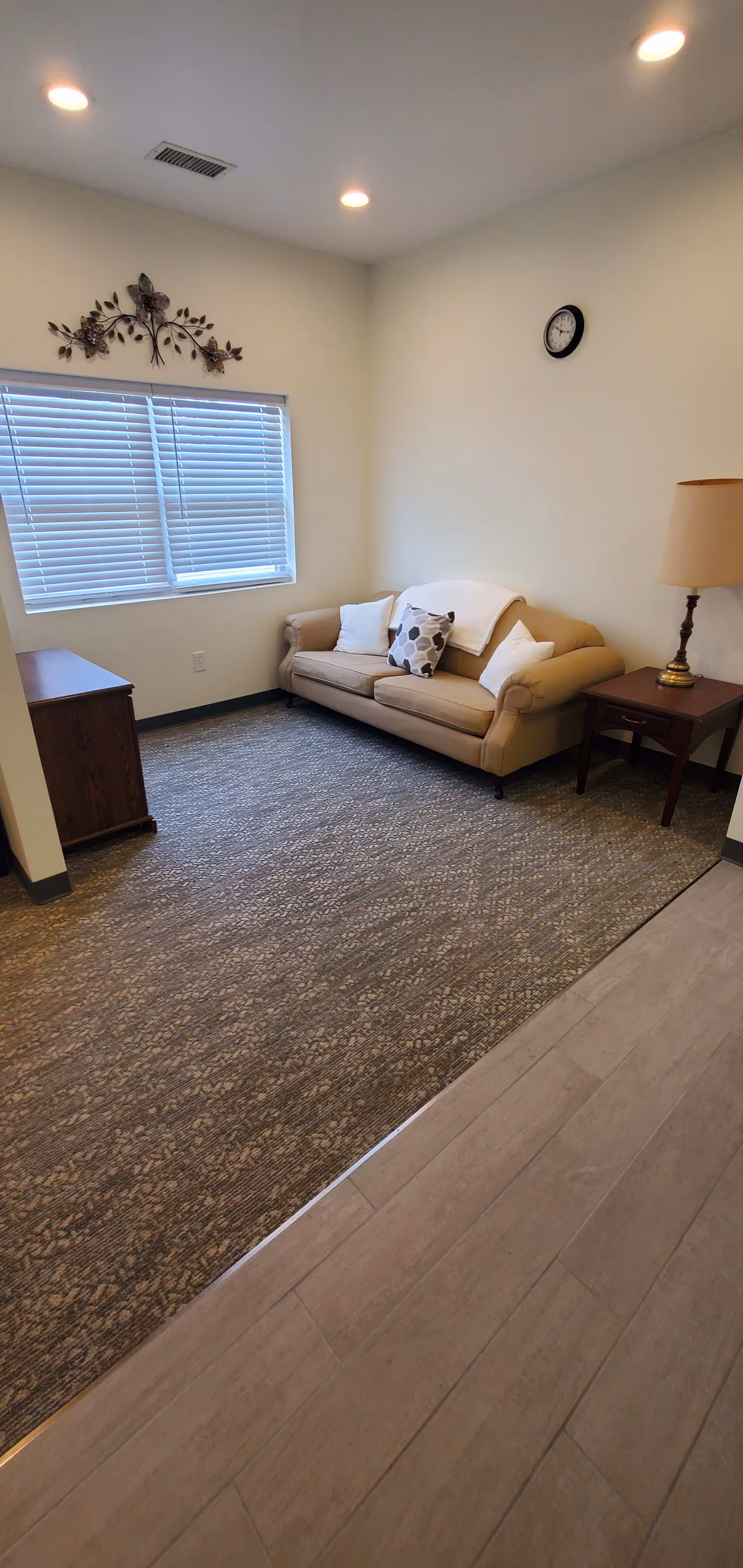 A small living area with a beige couch adorned with three pillows and a white throw blanket. Next to the couch is a wooden side table with a lamp. Across from the couch is a wooden cabinet. The room has a window with closed blinds, a decorative metal wall art above the window, a wall clock, recessed ceiling lights, and a carpeted floor transitioning to a tiled floor.