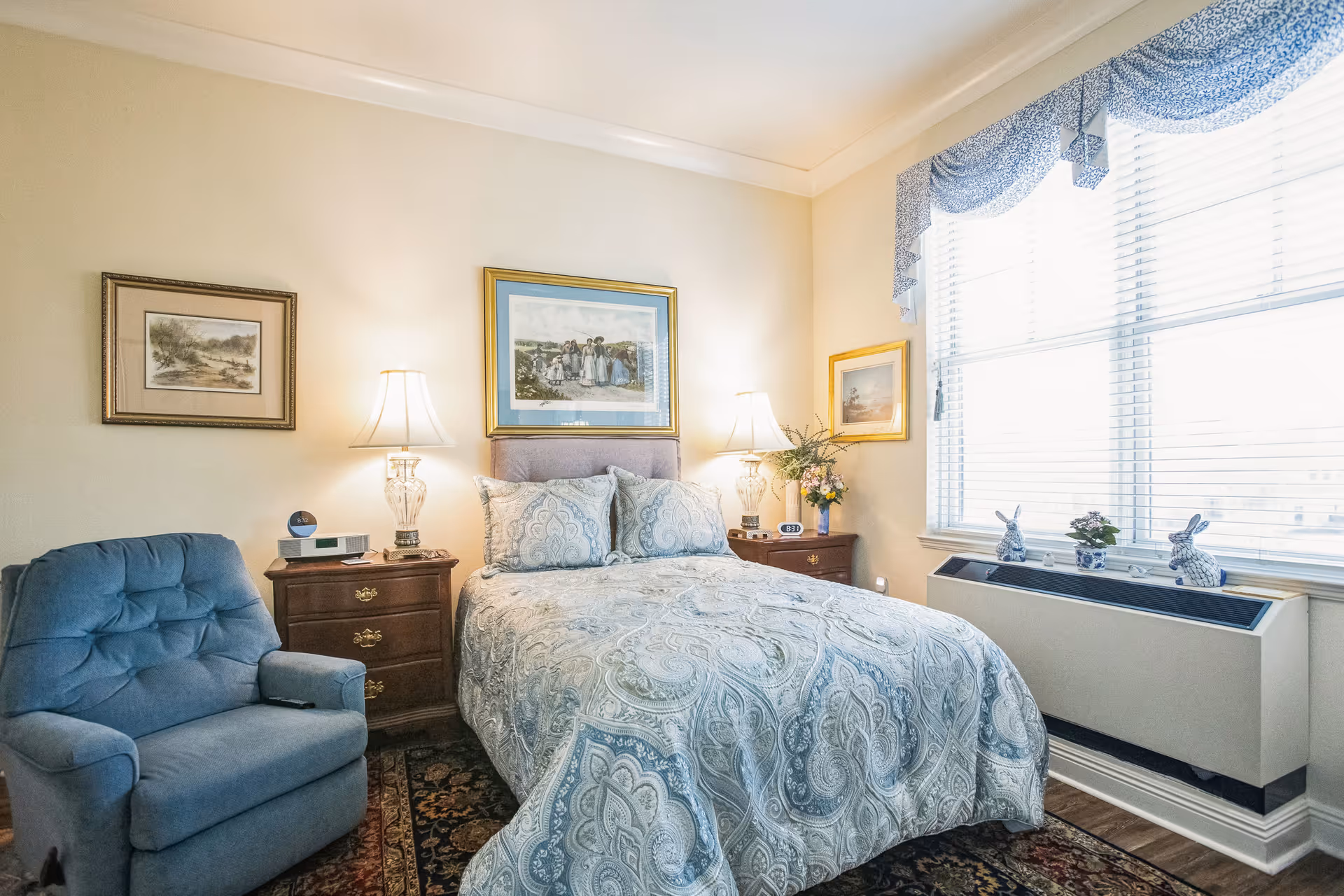 A cozy bedroom with a bed covered in a blue and white patterned comforter and matching pillows. There are two wooden nightstands on either side of the bed, each with a lamp and decorative items. A blue upholstered armchair is positioned to the left of the bed. The room has framed artwork on the walls and a large window with blue valance curtains, letting in natural light. Below the window are small decorative items including two ceramic rabbits and a small plant.