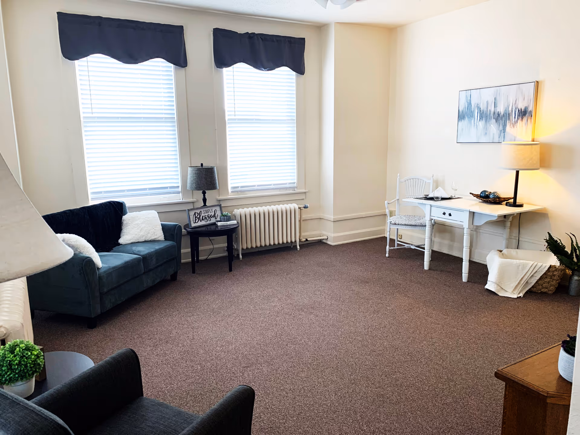 A cozy living room with two large windows covered by white blinds and dark valances. The room features a dark green sofa with white pillows, a small round side table with a lamp and decorative sign, a white desk with a chair, a table lamp, and decorative items. The carpet is brown, and the walls are light-colored with a piece of abstract art hanging above the desk.