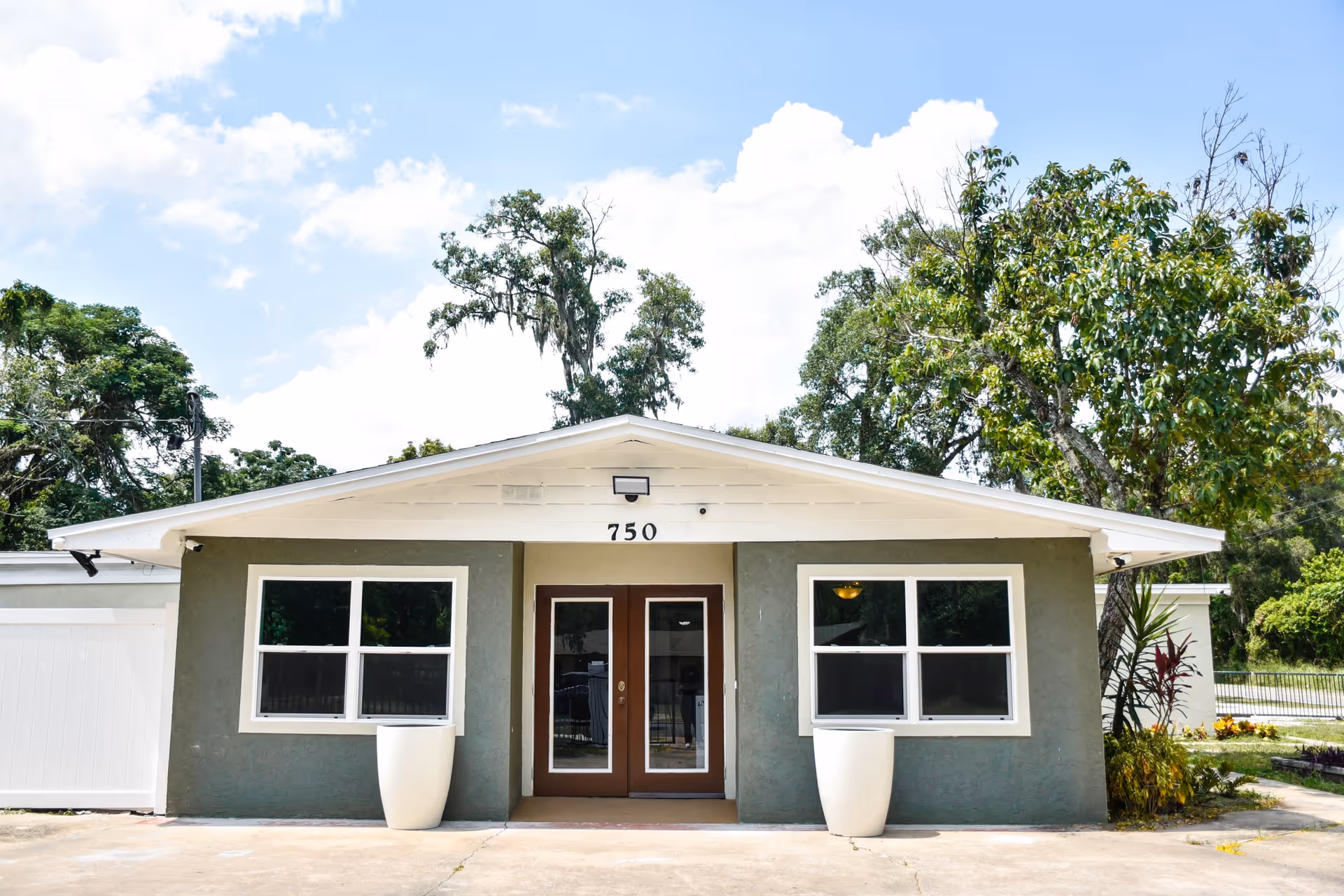 Front exterior view of a single-story building with gray walls, white trim, and a white roof. The building has two large windows on either side of a double glass door entrance with the number 750 above it. There are two large white planters in front of the windows and trees surrounding the building under a partly cloudy sky.