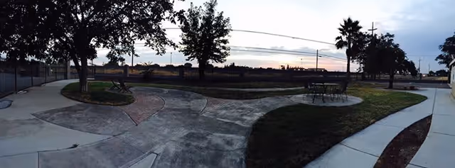 Outdoor courtyard with curved concrete walkways, benches and tables, trees, and a fenced grassy area at sunset.