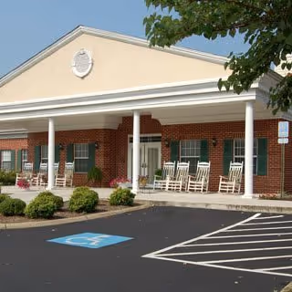 Front exterior of a single-story brick senior living building with a covered porch, white columns, rocking chairs, and accessible parking.