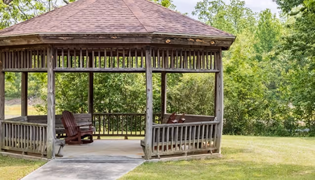 A wooden gazebo with a shingled roof situated in a grassy area surrounded by trees, featuring wooden benches and chairs inside.