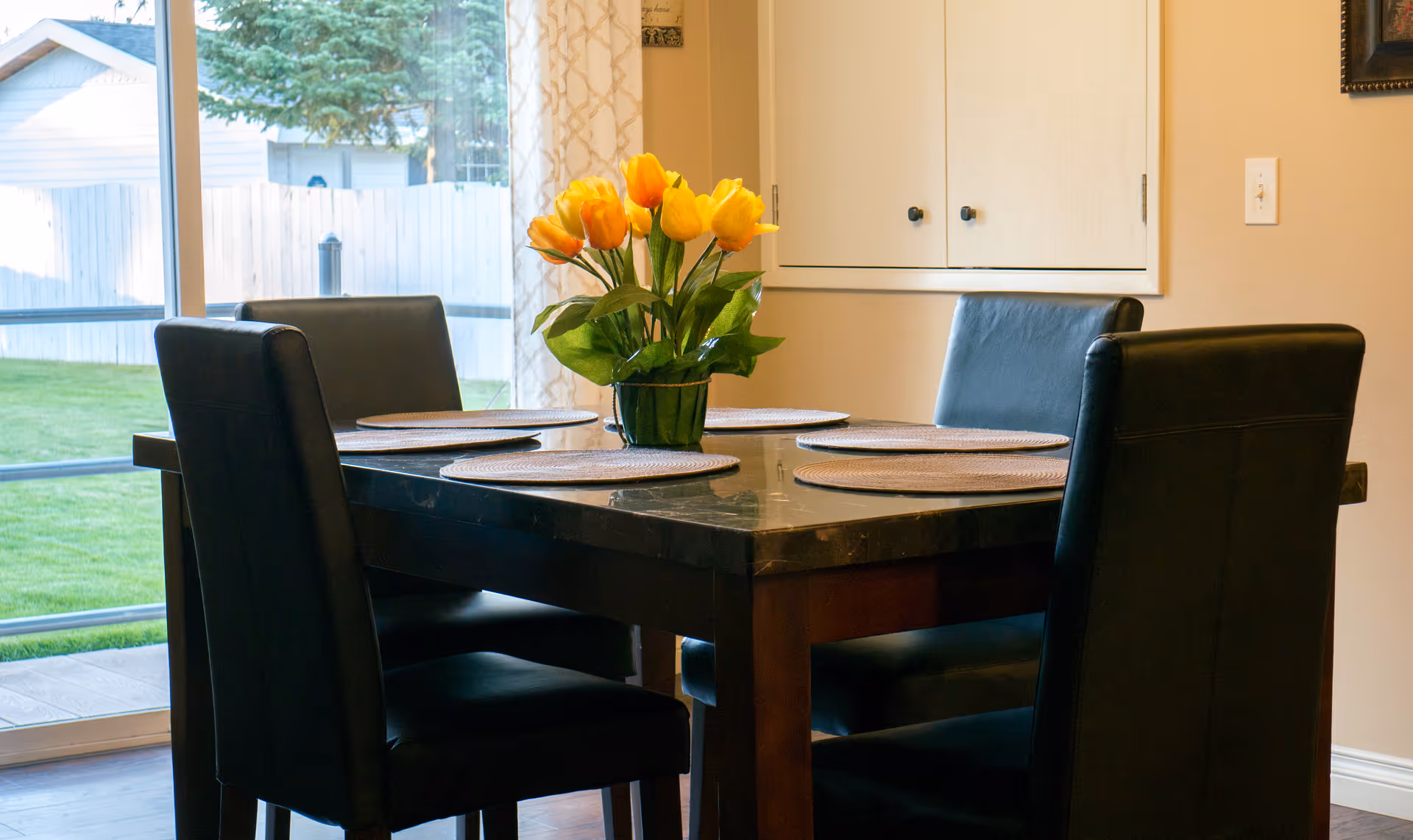 Dining room with a dark table set for four, black chairs and a vase of yellow tulips by a sliding glass door.