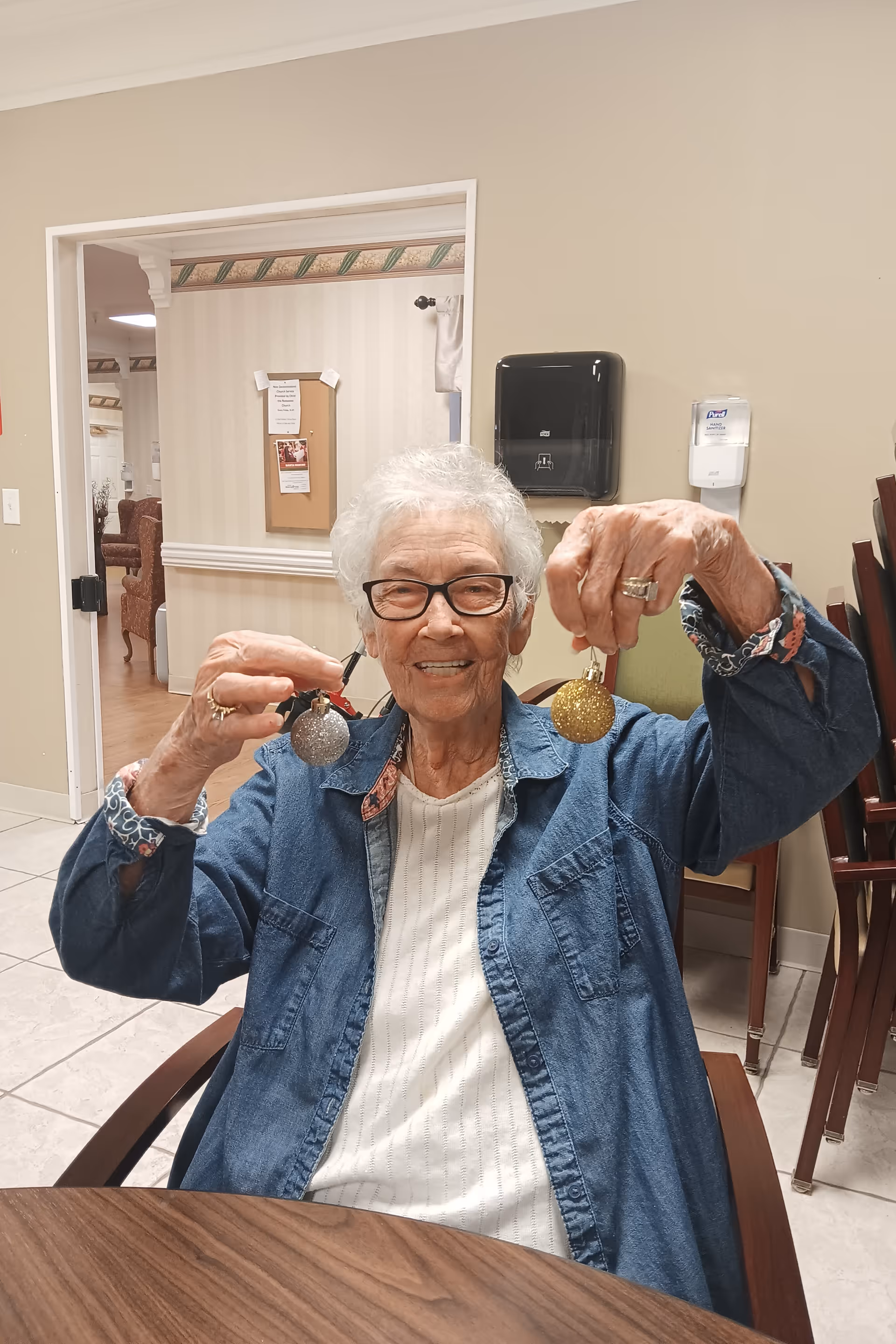 An elderly woman with white hair and glasses is sitting at a table indoors, smiling and holding up two Christmas ornaments, one silver and one gold. She is wearing a white top and a blue denim jacket. Behind her, there is a beige wall with a paper towel dispenser and a hand sanitizer dispenser, and some chairs stacked in the corner.