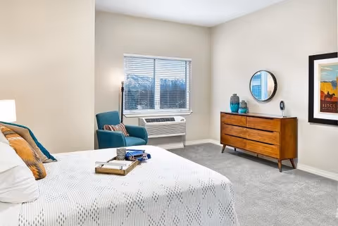 A bright and clean bedroom in an assisted living facility featuring a neatly made bed with white bedding and decorative pillows, a wooden dresser with a round mirror and decorative vases, a framed artwork on the wall, a teal armchair with a striped pillow, a floor lamp, and a window with blinds letting in natural light.