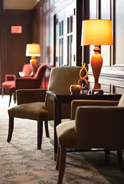 A warmly lit interior seating area with upholstered chairs, side tables, and table lamps along a carpeted corridor.