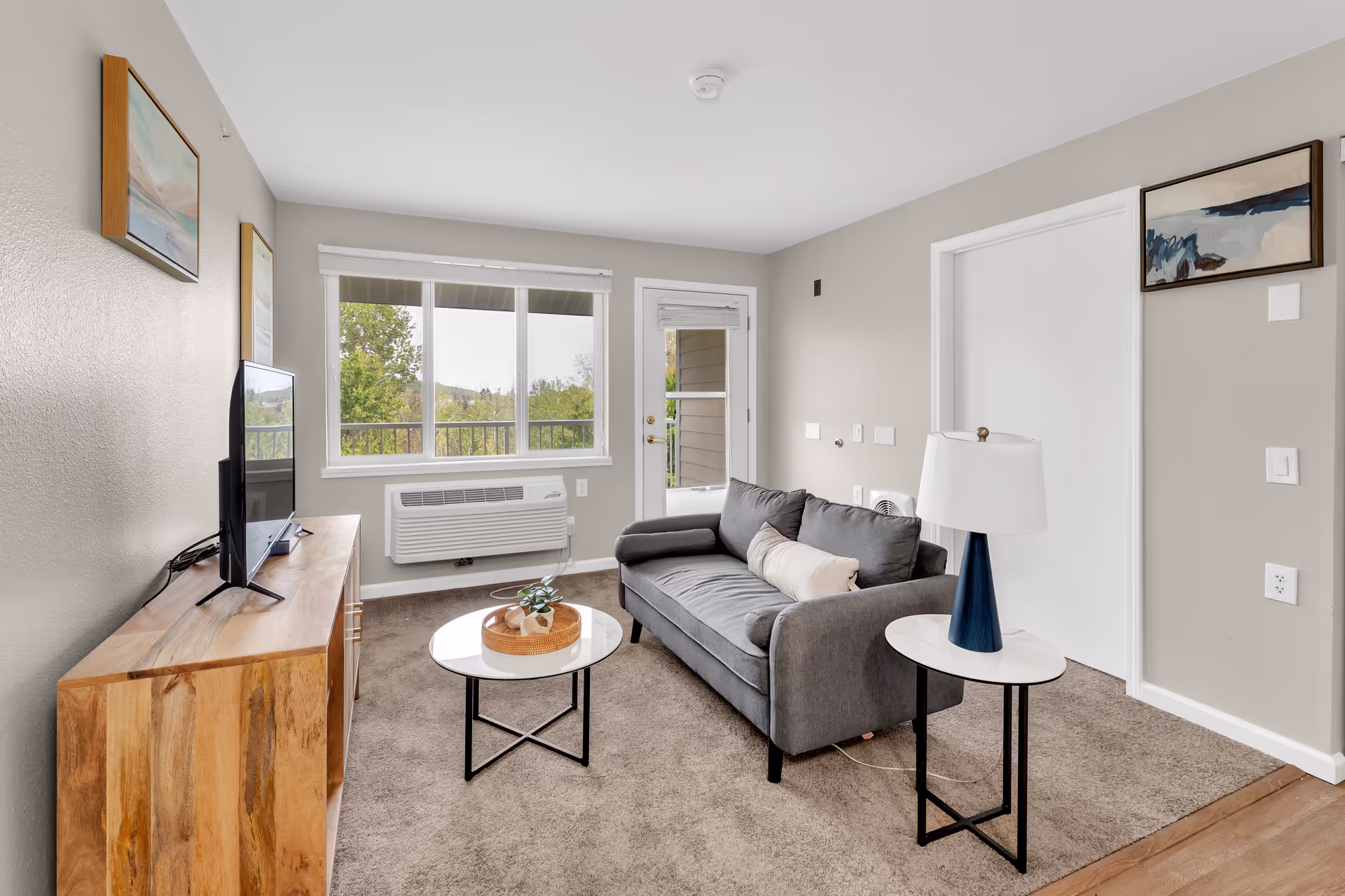A cozy living room with a gray sofa, a small round coffee table with a decorative tray, a side table with a lamp, a wooden TV stand with a flat-screen TV, and large windows showing greenery outside. The walls are light gray with framed artwork, and there is a door leading to an outdoor balcony.