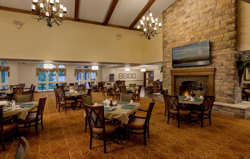 A spacious dining room with multiple tables covered in beige tablecloths and green placemats, set with cups and condiments. The room features a high ceiling with wooden beams and chandeliers, large windows with decorative valances, and a stone fireplace with a mounted flat-screen TV above it.