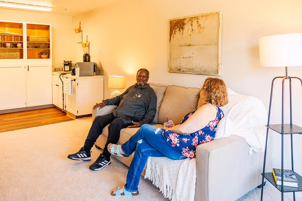 Two people sitting and conversing on a sofa in a bright living room with cabinets and a floor lamp.
